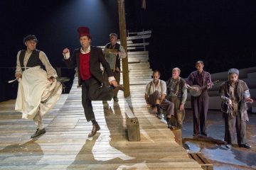 Näyttelijät esiintyvät laivaa muistuttavalla puisella lavasteella. Kaksi edessä tanssii energisesti, kun taas toiset istuvat tai seisovat taustalla, jotkut soittimien kanssa, vanhanaikaisiin, maalaismaisiin vaatteisiin pukeutuneina. Valaistus on dramaattinen.