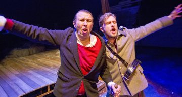 Kaksi miestä lavalla intohimoisesti laulamassa tai huutamassa, kädet ojennettuina. Toisella on punainen pusero ja musta takki, toisella harmaa sotilastyylinen takki, jossa on kamera. Puiset lankut ja hämärä valaistus viittaavat teatteriesitykseen.