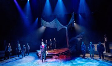 Dramaattisessa näyttämökohtauksessa, jossa on sininen valaistus, näyttelijät seisovat rikkoutunutta laivaa muistuttavan lavastuksen ympärillä. Keskeinen näyttelijä seisoo edessä valokeilassa, kun taas muut seisovat hajallaan ympäri näyttämöä, mikä luo intensiivisen tunnelman.