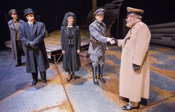 Viisi aikakauden pukuihin pukeutunutta näyttelijää seisoo näyttämöllä. Kaksi sotilaspukuista miestä kättelee, kun taas mustiin pukeutunut nainen ja kaksi juhlapukuista miestä tarkkailevat. Lavastuksessa on puinen lava ja vain vähän rekvisiittaa.