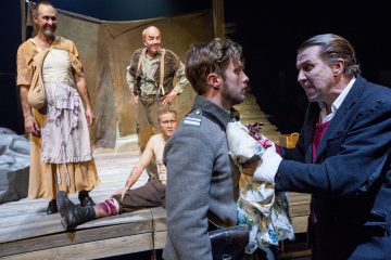 Neljä näyttelijää esiintyy puisella näyttämöllä, ja yksi heistä on sotilaspuvussa, ja vanhempi mies tarttuu häneen. Kaksi muuta katselee, toinen riekaleisessa mekossa ja toinen lattialla istuen, kaikki dramaattisissa, ilmeikkäissä asennoissa.