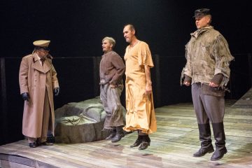 Neljä näyttelijää seisoo näyttämöllä: yhdellä on sotilastakki ja -lakki, toisella rähjäiset vaatteet, kolmas on paljain jaloin yksinkertaisessa oranssissa kaapussa ja neljäs on laastaripukuinen ja lippalakki päässä; he näyttävät olevan kesken esityksen.