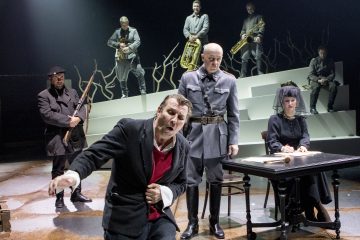 Mies puristaa rintaansa ja näyttää ahdistuneelta ankaran upseerin, pöydän ääressä istuvan naisen ja kolmen puhallinsoittimia soittavan muusikon edessä korotetulla korokkeella, jonka taustalla on paljaita oksia.