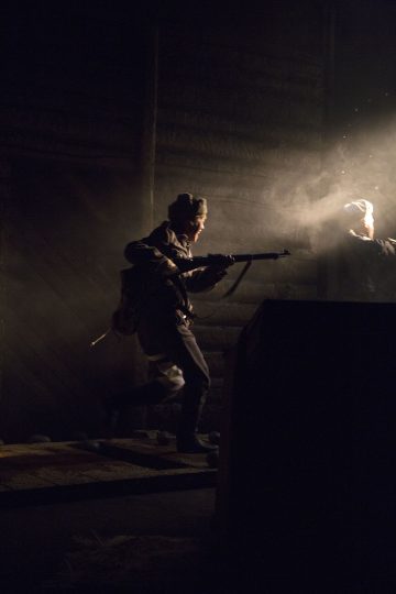 Kivääri kädessä oleva henkilö liikkuu varovasti hämärässä, savuisessa huoneessa, jonka oikeasta yläkulmasta tulvii valoa, joka valaisee osan hänen kasvoistaan ja vartalostaan.