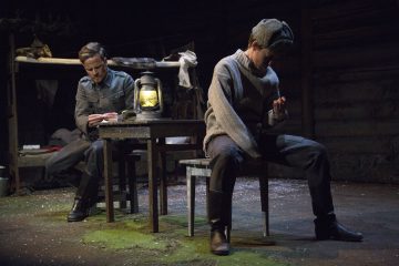 Kaksi miestä istuu hämärässä, maalaismaisessa huoneessa; toinen istuu pöydän ääressä ja kirjoittaa lyhdyn vieressä, toinen istuu jakkaralla ja katsoo alaspäin. Taustalla näkyy kerrossänky ja puiset seinät, jotka viittaavat maaseutu- tai sota-ajan ympäristöön.