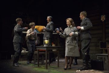 Viisi sotilasasuihin pukeutunutta näyttelijää esittää näyttämöllä vilkkaan kohtauksen, jossa yksi henkilö makaa ylimmällä punkalla, muut pitelevät juomia ja hymyilevät. Lavastus muistuttaa maalaismaista mökkiä, jonka keskellä on pöytä ja lyhty.