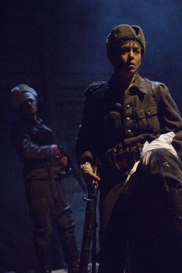 Kaksi sotilaspukuista henkilöä seisoo dramaattisessa valaistuksessa; edessä olevalla henkilöllä on turkislakki, hänellä on kivääri kädessään ja hän näyttää päättäväiseltä, kun taas toinen henkilö, jolla on kivääri, seisoo taustalla, osittain varjossa.