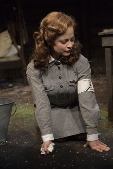 Ruskeahiuksinen nainen, jolla on harmaa univormu ja käsivarsinauha, polvistuu maahan ja puhdistaa liinalla. Lähistöllä on metallinen ämpäri ja hajallaan olevia roskia, ja taustalla on hämärä, maalaismainen ympäristö.