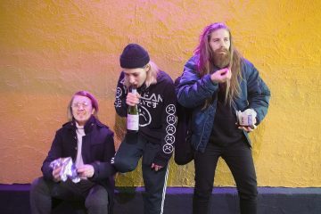 Kolme rennosti pukeutunutta ihmistä seisoo keltaista seinää vasten. Yksi istuu syömässä kääreestä, toinen juo pullosta, ja kolmas, pitkätukkainen, syö laatikosta, kaikki näyttävät rennoilta ja epämuodollisilta.