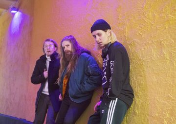 Kolme pitkätukkaista ja katupukuista nuorta aikuista poseeraa keltaista seinää vasten violetissa valaistuksessa ja katsoo kameraan. Yhdellä on silmälasit, toisella pipo, ja kaikilla on rennot, urbaanit vaatteet.