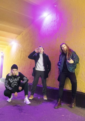 Kolme ihmistä poseeraa vaaleanpunaisessa valaistuksessa keltaisen seinän vieressä; yksi kyykistyy pipo ja graafinen collegepaita yllään, toinen seisoo käsi kasvoillaan ja kolmas seisoo pitkät hiukset ja tekee kädellään eleen.