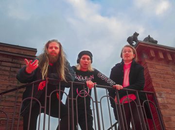 Kolme ihmistä seisoo mustan rauta-aidan takana tiiliparvekkeella ja katsoo alas ilmeikkäin kasvoin. Yhdellä on pitkät hiukset ja parta, toisella on pipo, kolmannella on silmälasit. Taivas on sininen ja pilvinen heidän yläpuolellaan.