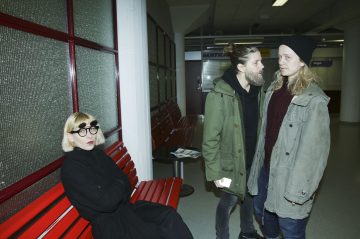 Mustiin vaatteisiin ja pyöreisiin silmälaseihin pukeutunut nainen istuu yksin punaisella penkillä ja katsoo kameraan, kun taas kaksi vihreisiin takkeihin pukeutunutta ihmistä seisoo lähekkäin lähekkäin ja puhuu käytävällä.