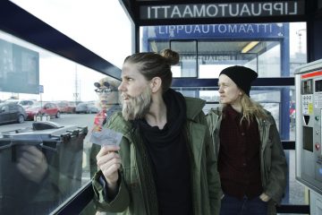 Kaksi ihmistä seisoo lasiseinäisen bussisuojan sisällä. Toinen pitää korttia kädessään ja katsoo ulos, kun taas toinen katsoo toiseen suuntaan. Kohtaus on urbaani, ja näkyvissä on autoja ja lippuautomaatti. Molemmilla on rennot takit ja hatut.