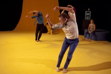 Kaksi paljasjalkaista, rennosti pukeutunutta tanssijaa tekee ilmeikkäitä liikkeitä kirkkaan keltaisella lavalla, kun taustalla istuva nainen tarkkailee heidän esitystään.