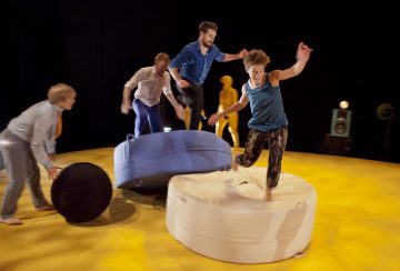 Neljä ihmistä hyppii ja tasapainoilee tarmokkaasti suurilla pyöreillä tyynyillä keltaisella lavalla, ja taustalla on abstraktia rekvisiittaa ja kaiutin.