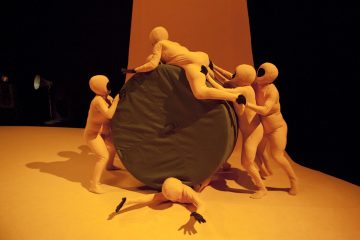 Kuusi ihmistä keltaisissa kokokuvapuvuissa on vuorovaikutuksessa suuren, pyöreän, vihreän esineen kanssa keltaisella näyttämöllä luoden abstraktin, surrealistisen kohtauksen dramaattisessa valaistuksessa.