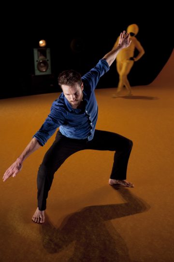 Siniseen paitaan ja mustiin housuihin pukeutunut paljasjalkainen mies tanssii dynaamisessa tanssipoosissa keltaisella lavalla, kun taustalla seisoo keltaiseen bodyyn pukeutunut henkilö.