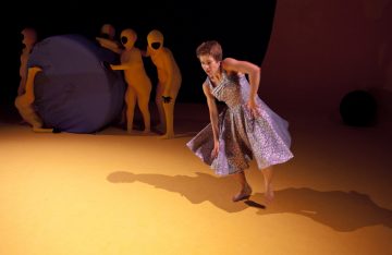 Kuviomekkoon pukeutunut paljasjalkainen nainen tanssii keltaisella näyttämöllä, kun taas keltaisiin kokovartalopukuihin pukeutuneet ihmiset liikuttavat suurta sinistä palloa taustalla.