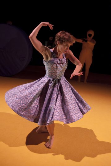 Paljasjalkainen tanssija, jolla on kuvioitu sininen mekko, poseeraa keltaisella lavalla ilmeikkäästi, toinen käsi koholla ja toinen taivutettuna. Taustalla hämärässä valaistuksessa seisoo beigeen bodyyn pukeutunut hahmo.