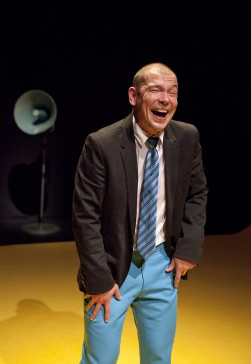 Mies, jolla on puvun takki, raidallinen solmio ja kirkkaansiniset housut, seisoo nauraen kädet polvillaan. Hänen takanaan näkyy suuri sininen megafoni tummaa taustaa vasten.