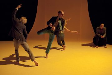 Neljä pukuihin pukeutunutta paljasjalkaista ihmistä esittää ilmeikkäitä tanssiasentoja lämpimässä, keltaisessa näyttämövalaistuksessa. Kolme seisoo tai liikkuu tarmokkaasti, kun taas yksi istuu oikealla ja katselee muita. Tausta on minimalistinen ja oranssin sävyinen.