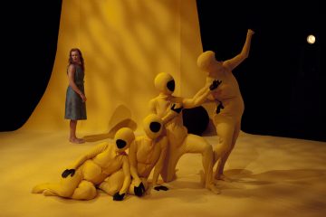 Siniseen mekkoon pukeutunut nainen seisoo keltaisella näyttämöllä ja katselee neljää keltaisiin kokovartalopukuihin pukeutunutta hahmoa, joilla on mustat hanskat ja kasvosuojukset ja jotka poseeraavat ja ovat vuorovaikutuksessa hänen edessään.