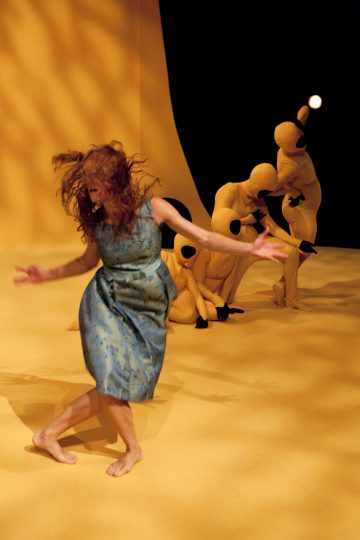 Siniseen mekkoon pukeutunut paljasjalkainen nainen tanssii keltaisella lattialla, ja taustalla poseeraa dramaattisesti keltaista taustaa vasten useita keltaisiin kokovartalopukuihin pukeutuneita ihmisiä.