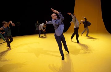 Kuusi tanssijaa tekee ilmeikkäitä liikkeitä kirkkaan keltaisella näyttämöllä, jossa on kaareva tausta, ja jokainen tanssija tekee ainutlaatuisen poseerauksen. Tausta on tumma, mikä korostaa eloisaa lattiaa ja heidän dynaamisia asentojaan.