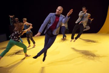 Ryhmä värikkäisiin asuihin pukeutuneita paljasjalkaisia tanssijoita hyppii ja liikkuu energisesti keltaisella näyttämöllä kesken esityksen ilmeikkäin elein ja dynaamisin liikkein.