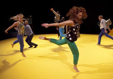 Kuusi ihmistä tanssii tarmokkaasti paljain jaloin kirkkaan keltaisella lattialla, ja dramaattiset liikkeet ja ilmeikkäät asennot on kuvattu kesken toiminnan mustaa taustaa vasten.