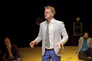 Vaaleaan pukuun ja solmioon pukeutunut mies seisoo näyttämön keskellä ja tekee ilmeikkäitä ilmeitä ja eleitä. Hänen takanaan keltaisella lattialla istuu kaksi muuta pukumiestä, joiden kaiuttimet näkyvät hämärässä taustalla.