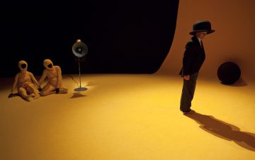 Mustaan pukuun ja suureen hattuun pukeutunut henkilö seisoo keltaisella lavalla ja heittää varjon. Kaksi keltaisiin bodyihin pukeutunutta hahmoa istuu lattialla kaiuttimen vieressä, ja taustalla on musta pallo. Kohtaus on dramaattisesti valaistu.