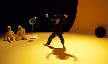 Pukuun ja keilahattuun pukeutunut henkilö tanssii paljain jaloin elinvoimaisen keltaisella lattialla ja heittää suuren varjon. Kaksi keltaisiin bodyihin pukeutunutta hahmoa istuu kaiuttimen ääressä taustalla keltaista ja mustaa taustaa vasten.