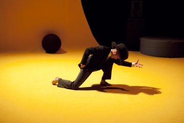 Tummaan pukuun ja leveälieriseen hattuun pukeutunut henkilö polvistuu kirkkaan keltaiselle lattialle ja ojentaa yhden kätensä dramaattisesti eteenpäin varjoa heittäen. Taustalla on abstrakteja, tummia muotoja.