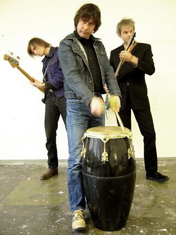 Kolme miestä soittaa musiikkia; yksi soittaa bassokitaraa, toinen sähkökitaraa, ja edessä oleva soittaa kongarumpua. He seisovat sisätiloissa betonilattialla vaaleaa seinää vasten.