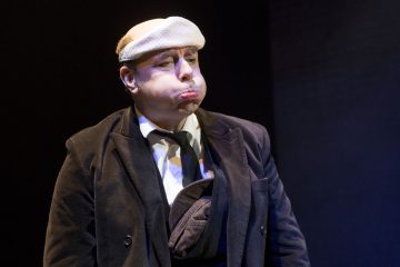 Pukuun ja solmioon pukeutunut mies, jonka pään ympärille on kiedottu valkoinen liina, paisuttaa dramaattisesti poskiaan ja seisoo näyttämövalaistuksessa tummaa taustaa vasten.