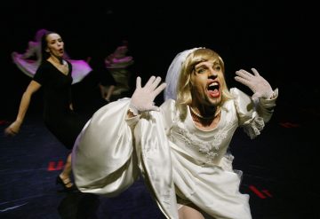 Dramaattiseen valkoiseen morsiuspukuun ja hanskoihin pukeutunut esiintyjä elehtii ilmeikkäästi lavalla, kun taas toinen mustiin pukeutunut henkilö tanssii taustalla tummalla taustalla.