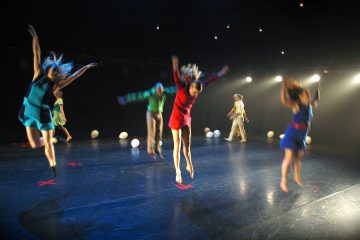 Viisi värikkäisiin asuihin pukeutunutta tanssijaa hyppii energisesti hämärästi valaistulla näyttämöllä, jonka lattialle on ripoteltu valkoisia palloja ja jonka taustalla loistavat näyttämövalot.