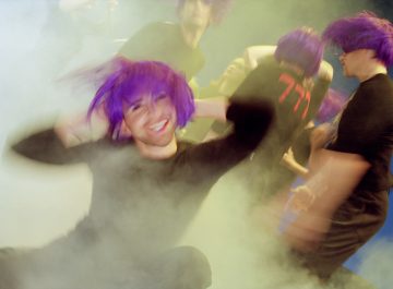 Ihmiset, joilla on eloisat violetit peruukit ja mustat asusteet, tanssivat energisesti pyörteisen savun ja värikkäiden valojen keskellä luoden vilkkaan, surrealistisen ja hieman epäselvän kohtauksen.