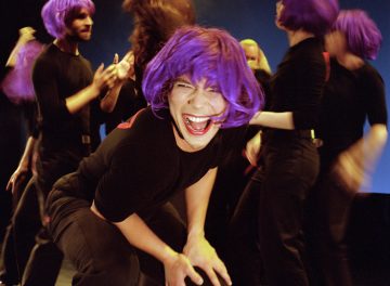 Ryhmä ihmisiä, joilla on kirkkaan violetit peruukit ja mustat asut, esiintyy energisesti lavalla, ja yksi henkilö etualalla nauraa ja kyyristyy kohti kameraa.