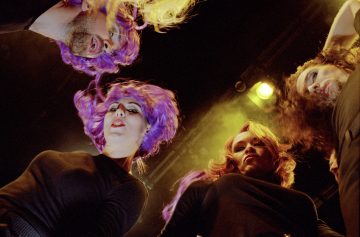 Neljä ihmistä, joilla on kirkkaan violetit ja vaaleanpunaiset peruukit ja mustat topit, seisovat piirissä ja katsovat kameraan. Dramaattinen valaistus ja näyttämötehosteet luovat teatteritunnelman.