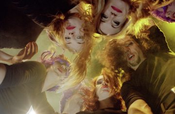 Kuuden dramaattisesti meikatun ja värikkäitä hiuksia kantavan ihmisen ryhmä nojaa tiiviisti yhteen ja katsoo kameraan päin ympyrän muotoisesti dramaattisessa valaistuksessa.