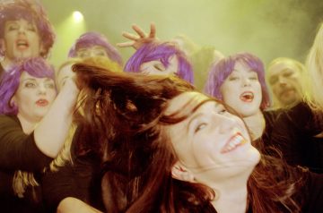 Iloinen ihmisjoukko, monilla kirkkaan violetit peruukit ja mustat vaatteet, juhlii yhdessä, kun yksi henkilö edessä kääntää pitkät ruskeat hiuksensa ja hymyilee kirkkaasti. Taustalla on näyttämön valojen hämärtämä valo.