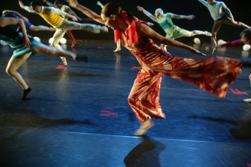 Ryhmä tanssijoita esiintyy tummalla lavalla, etualalla punaisessa mekossa oleva nainen, joka ojentaa kättään ja jalkaansa. Muut tanssijat ovat liikkeen sumentamia, mikä osoittaa energistä liikettä ja dynaamista koreografiaa.