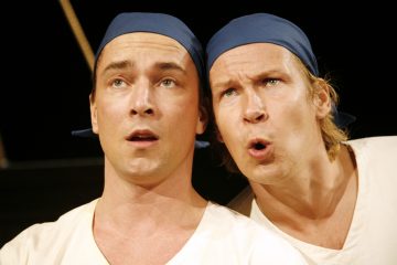 Kaksi miestä, joilla on siniset kaulaliinat ja valkoiset paidat, katsovat tarkkaavaisesti eteenpäin; toisella on yllättynyt ilme, kun taas toinen näyttää puhuvan tai reagoivan dramaattisesti.