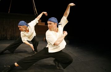 Kaksi esiintyjää valkoisissa paidoissa, mustissa housuissa ja sinisissä kaulaliinoissa poseeraavat dynaamisesti pimeällä näyttämöllä ja ojentavat kumpikin kätensä intensiivisesti ja keskittyneesti. Tausta on hämärästi valaistu, ja rekvisiittaa näkyy vain vähän.