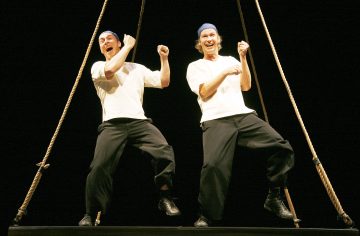 Kaksi esiintyjää, joilla on valkoiset paidat, mustat housut, siniset hatut ja mustat kengät, seisovat lähekkäin ripustetulla alustalla hymyillen ja elehtien, ja paksut köydet kehystävät heitä tummaa taustaa vasten.