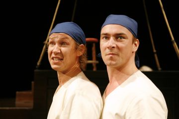 Lavalla seisoo kaksi miestä, joilla on siniset kaulaliinat ja valkoiset paidat, ja molemmilla on intensiiviset ja ilmeikkäät kasvonilmeet. Taustalla on tumma tausta, jossa on köysiä ja puinen jakkara.