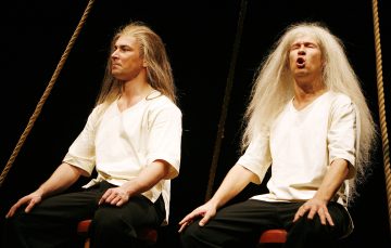 Kaksi pitkät, valkoiset hiukset omaavaa ihmistä istuu keinuilla valkoiset paidat ja mustat housut yllään tummaa taustaa vasten; toinen näyttää vakavalta, kun taas toisella on suu auki ikään kuin laulaisi tai puhuisi.